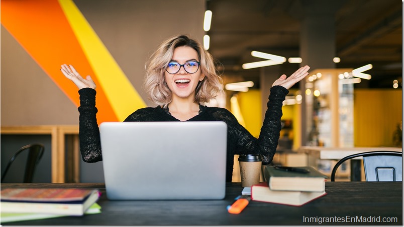 funny happy excited young pretty woman sitting at table in black shirt working on laptop in co-working office, wearing glasses